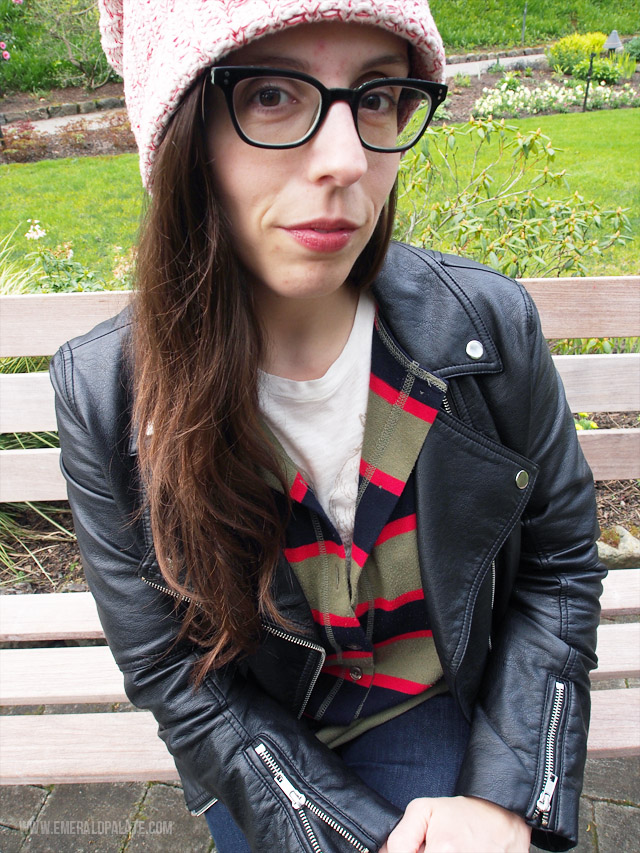 A closeup of woman wearing slouchy knit beanie, big black frame glasses, striped vintage henley rugby shirt, and a faux leather jacket.