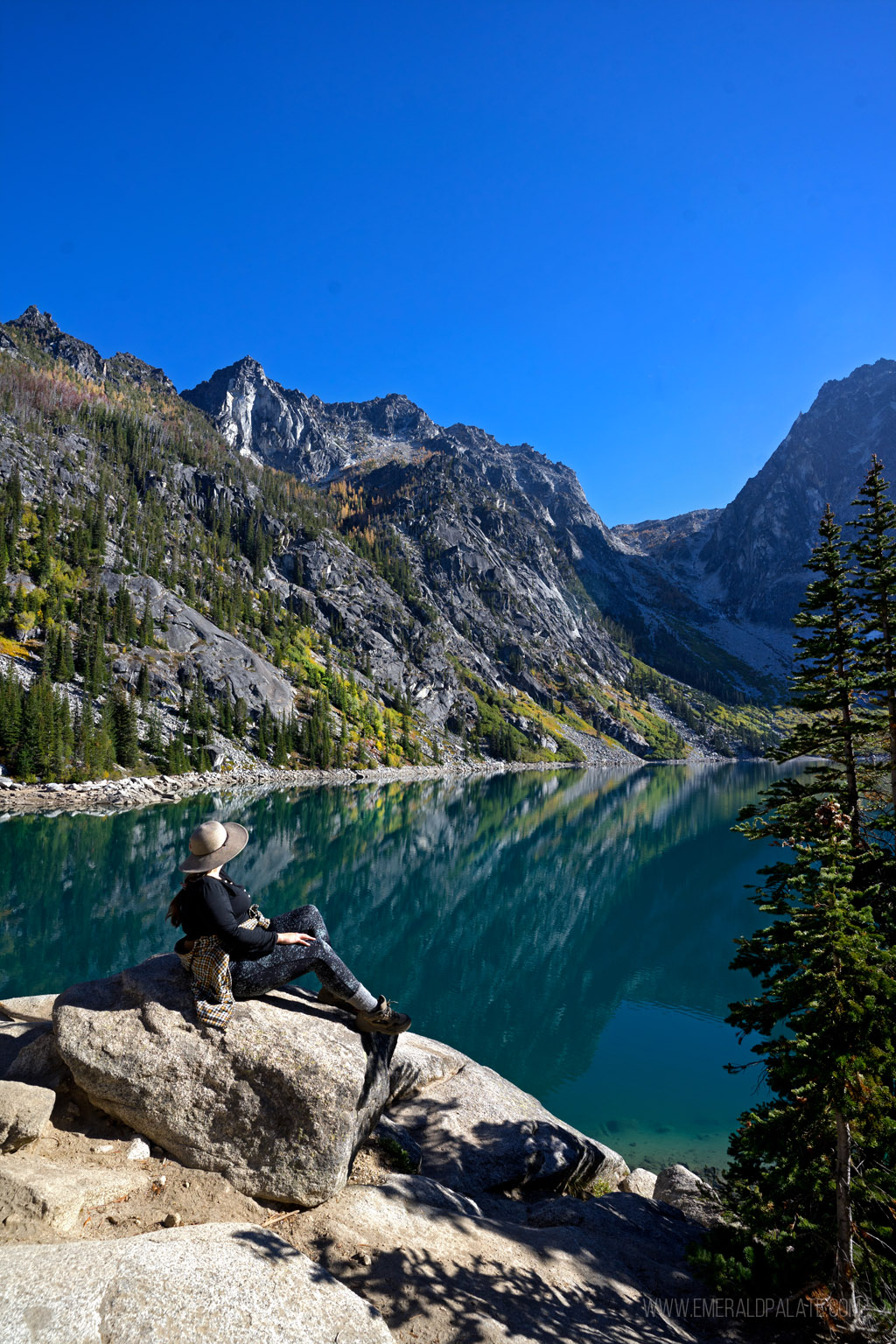 woman sitting on rock overlooking turquoise alpine lake dotted with larches