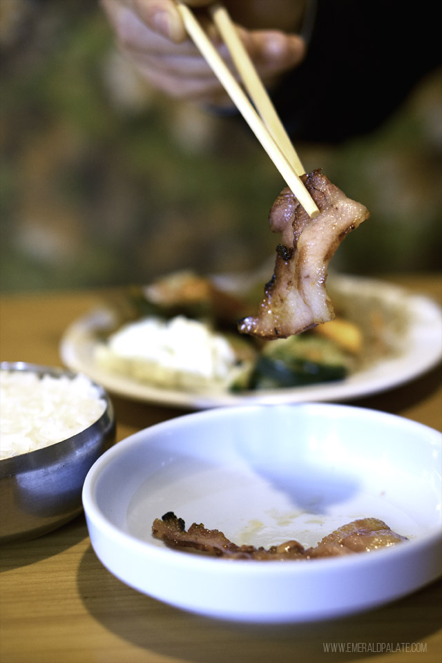 Person picking up pork jowl with chopsticks, which can be found at Cham Gardens, a restaurant in Lakewood, WA
