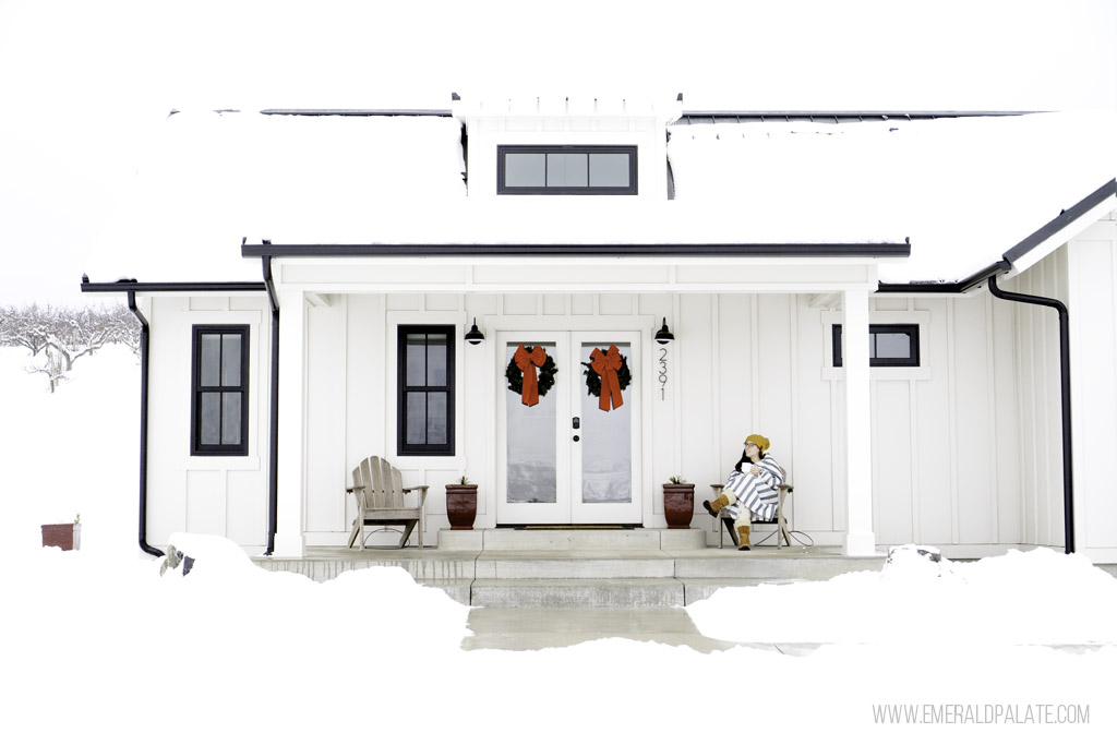 Place to stay in Lake Chelan covered in snow during Christmas time with wreaths on the door and a woman sitting on the porch with a blanket and beanie drinking coffee