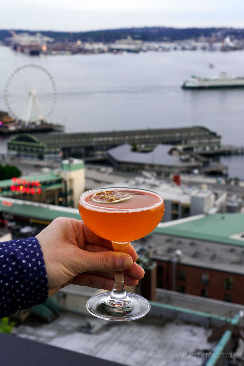 person holding a cocktail over the ledge of a Seattle rooftop bar with the Seattle waterfront in the background