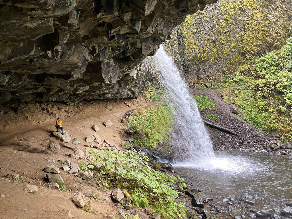 panoramic of a woman sitting in a cave underneath waterfall
