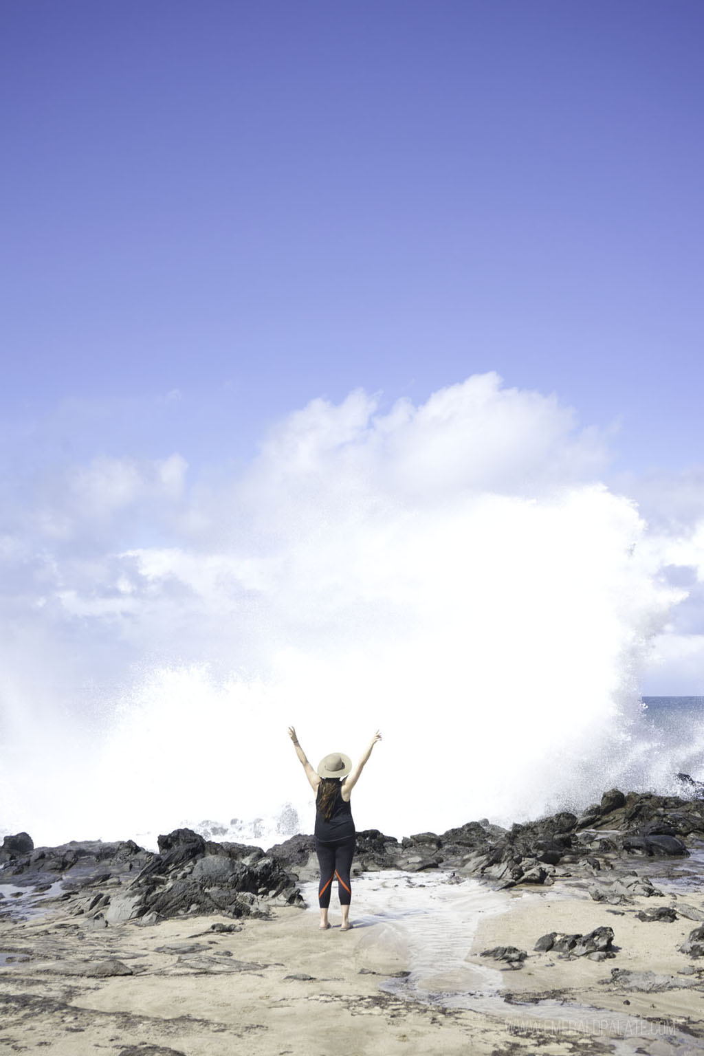 woman raising arms in celebration as a huge wave crashes on the rocks in front of her in Maui