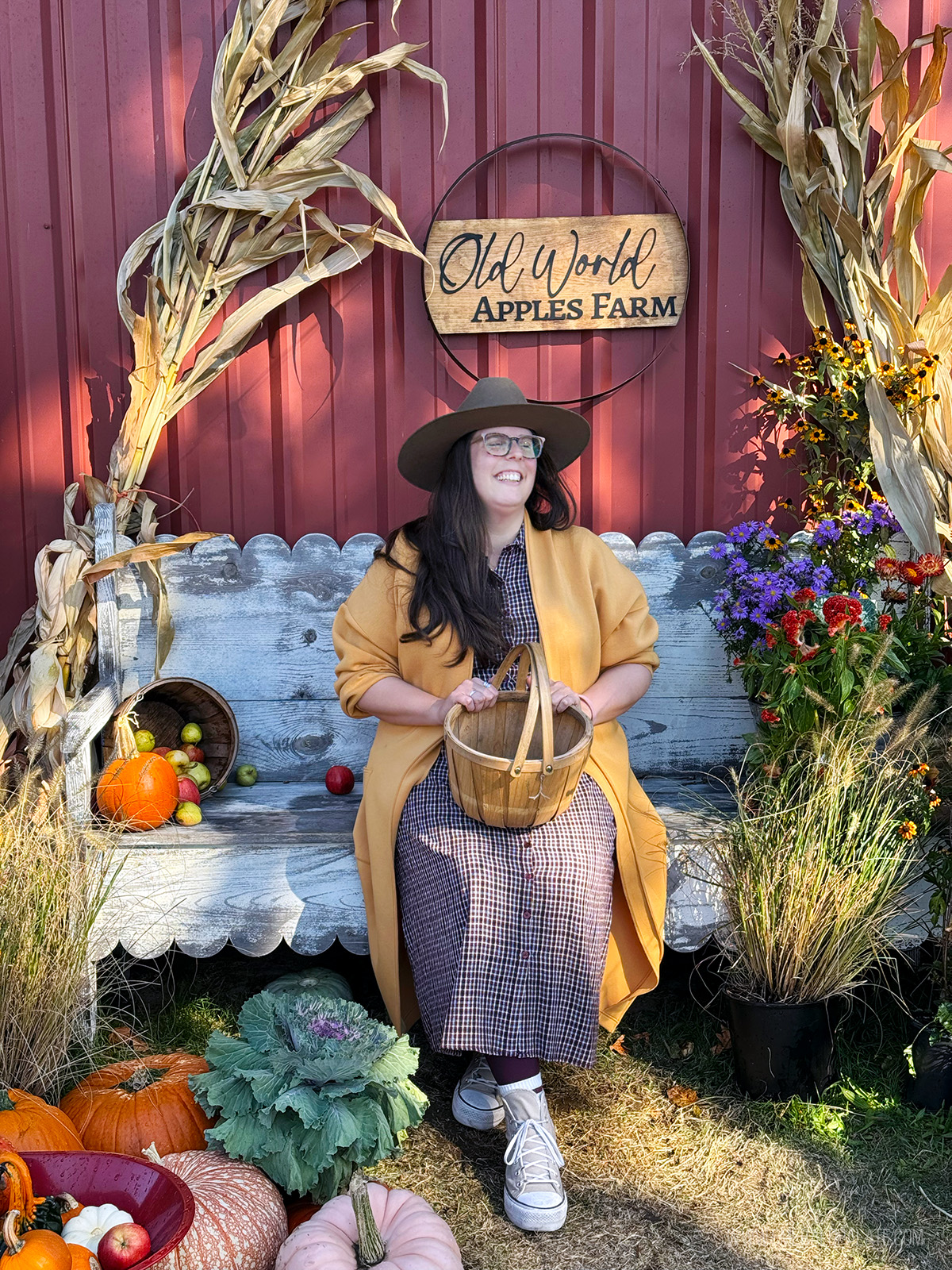 woman sitting in front of a fall photo set with corn husk, pumpkins, and flowers in front of a barn