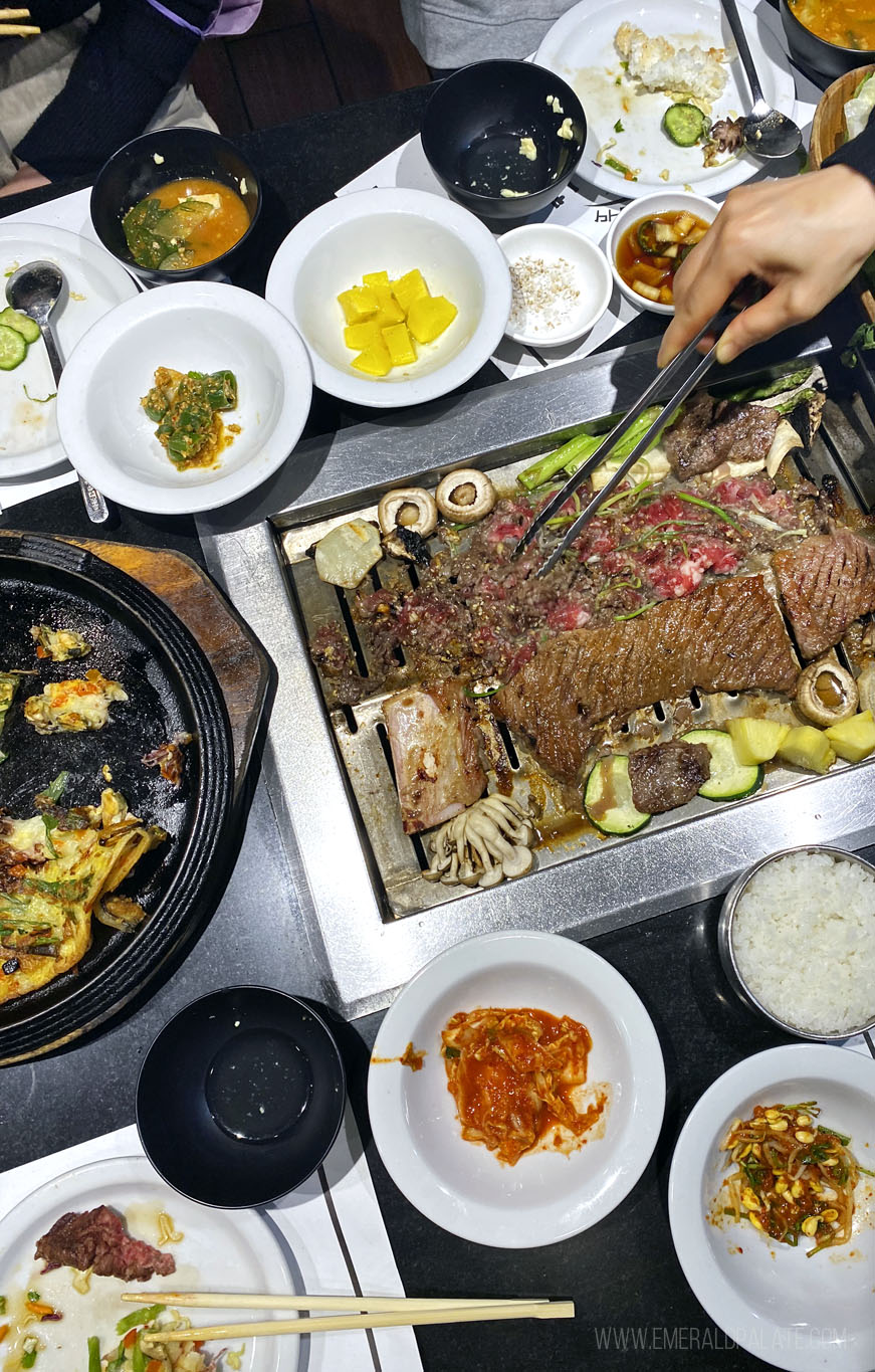 person grilling meat at a Korean BBQ restaurant table full of Korean food