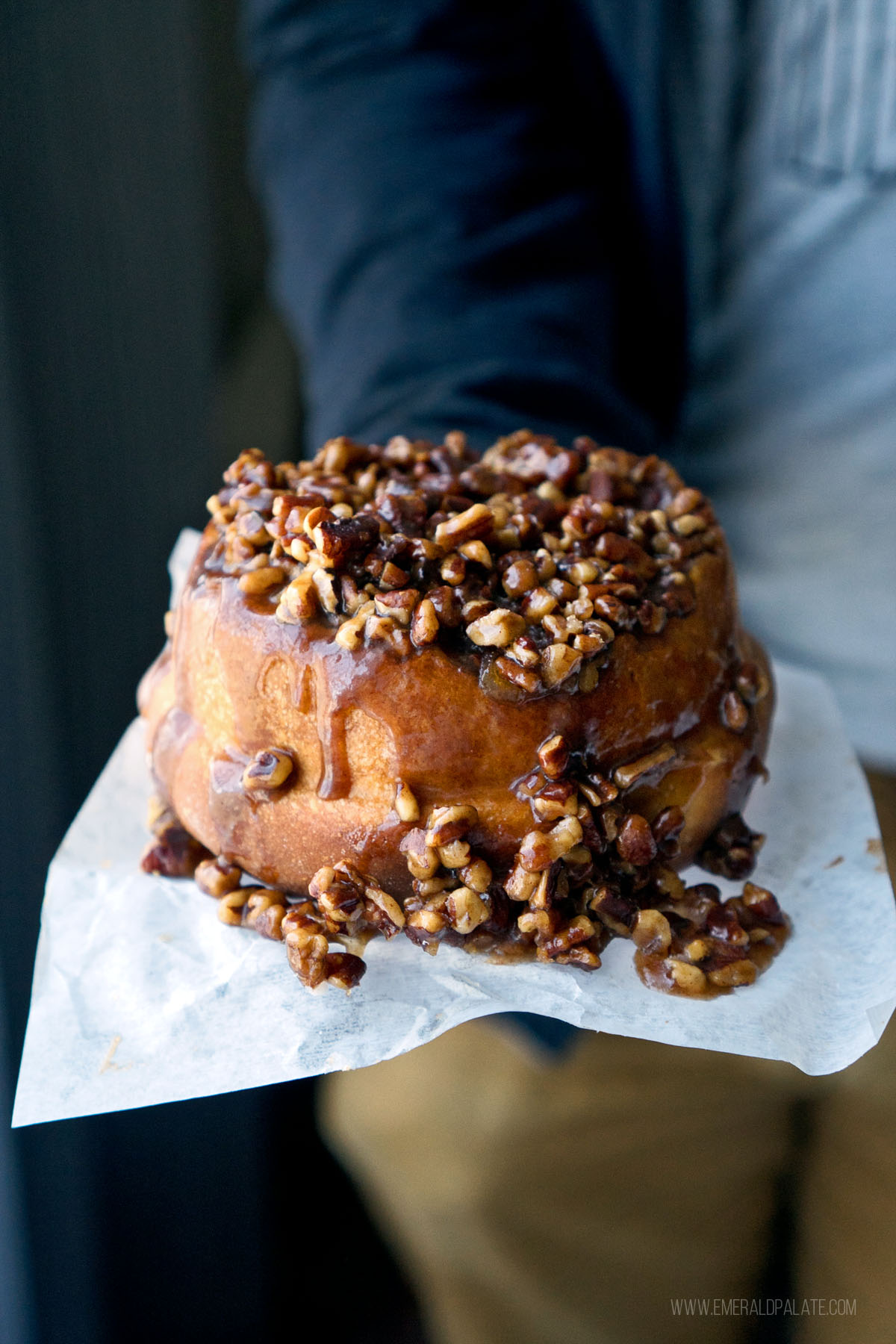 person holding a sticky cinnamon bun overflowing with candied pecans