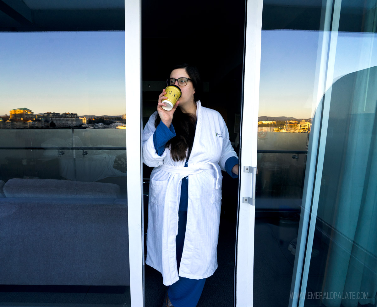 woman enjoying coffee in a robe from the terrace of a Victoria hotel with the waterfront reflecting in the glass at sunset