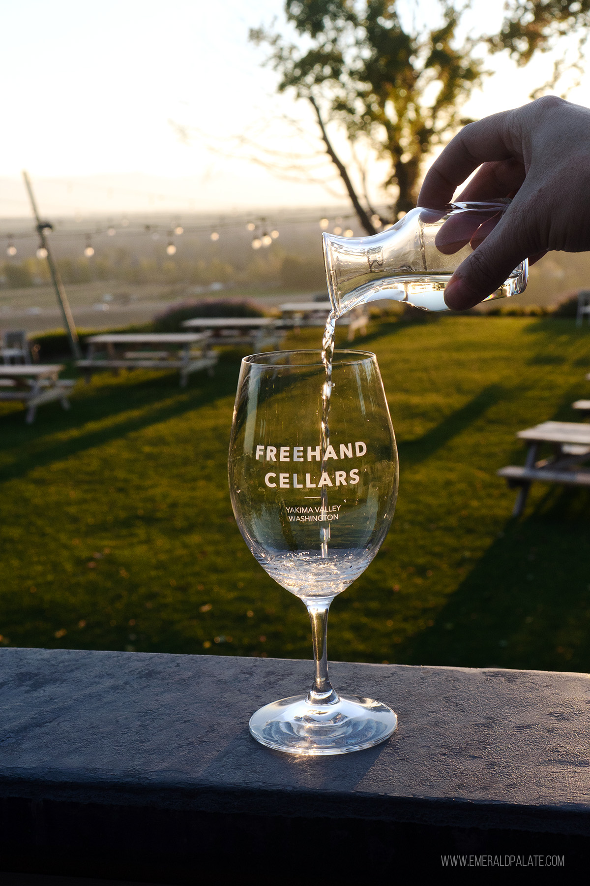 person pouring white wine into a glass with sunset views behind it