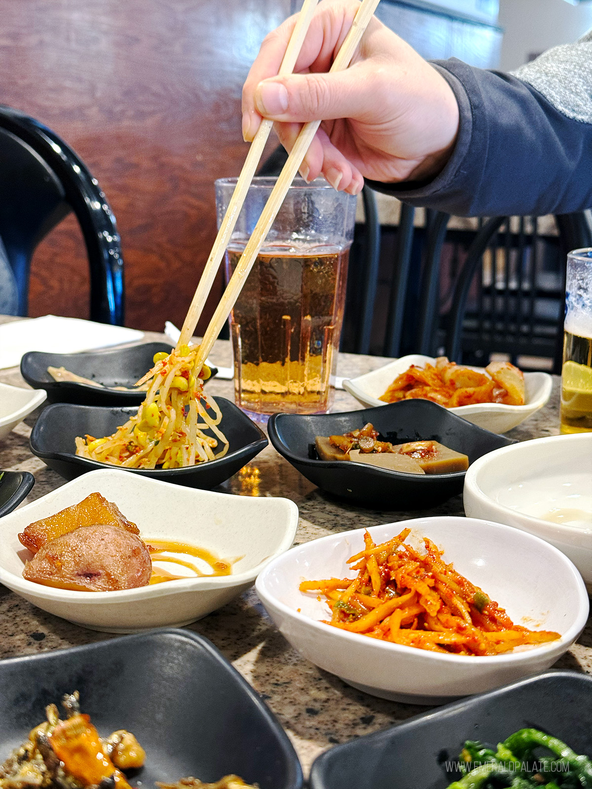 person picking up bean sprouts with chopped sticks among an array of Korean banchan