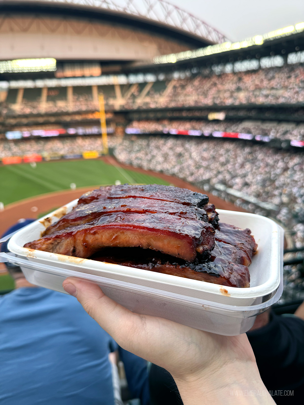 Person holding a plate of ribs in front of a baseball field at T Mobile Park